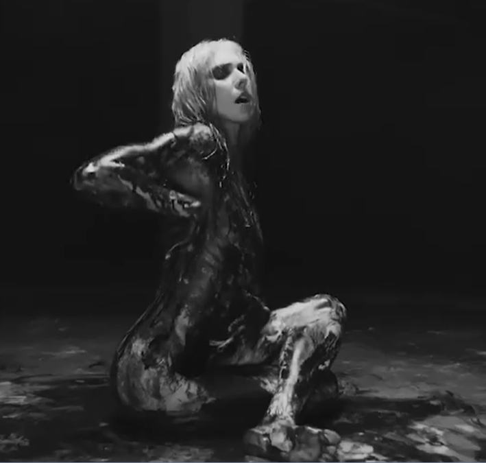 Photographie en noir et blanc d'une personne assise au sol, couverte de peinture sombre.