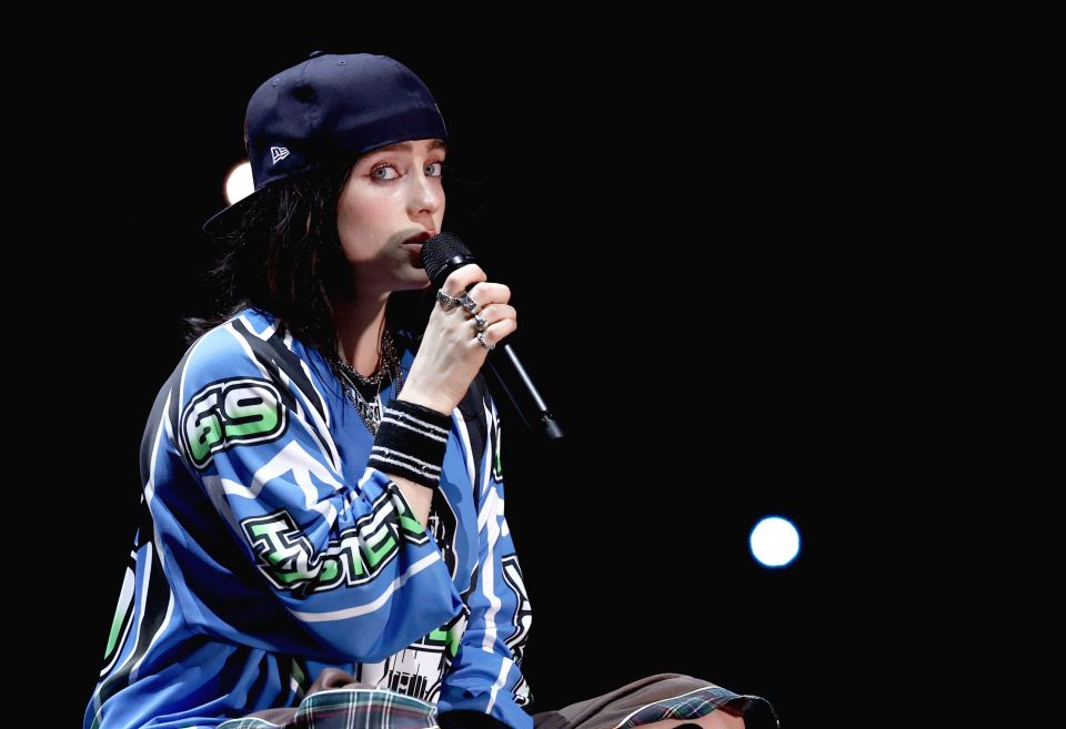 Billie Eilish performing onstage in a blue and white patterned shirt and a dark baseball cap.